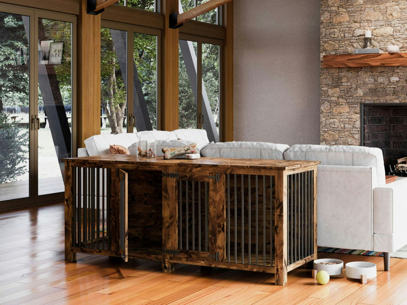 A Double Dog Kennel - Tuscany by James+James sits against the back of a white sectional sofa in a living room. The room features large windows with a scenic view, a stone fireplace, and various decor items. A stuffed toy and ball are on the hardwood floor nearby.