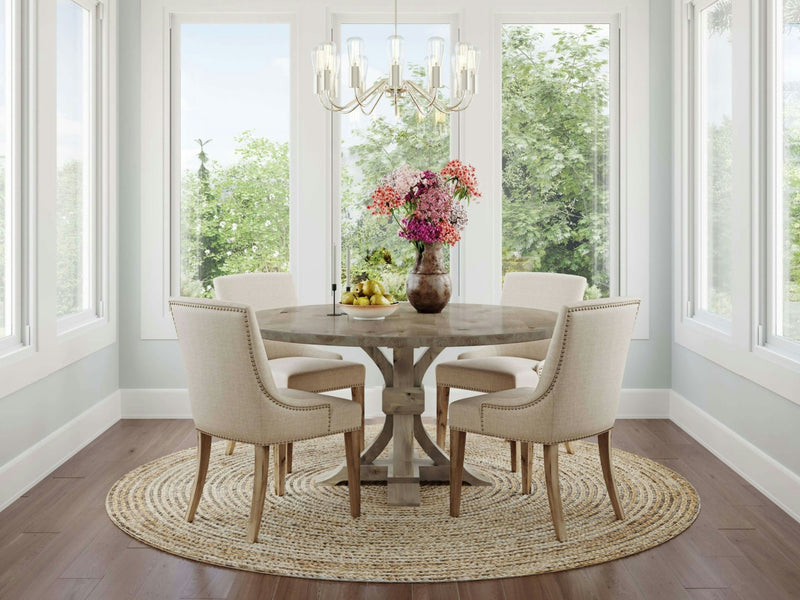 A bright dining room with large windows, allowing ample natural light. It features the Violet Round Dining Table in Barn Wood by James+James, surrounded by four cream-colored chairs. The table holds a vase with pink and white flowers and a bowl of lemons. A chandelier hangs above, and a wicker rug lies beneath.