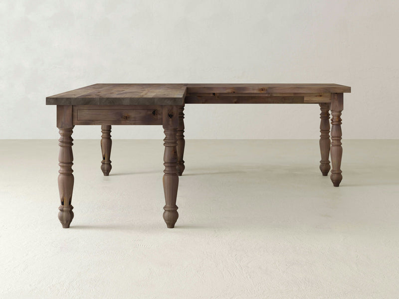 The Turned Leg Corner Desk - Barn Wood from James+James features an extended leaf on one side, a rustic finish, and intricately carved legs. It is set against a plain, light-colored wall and floor.