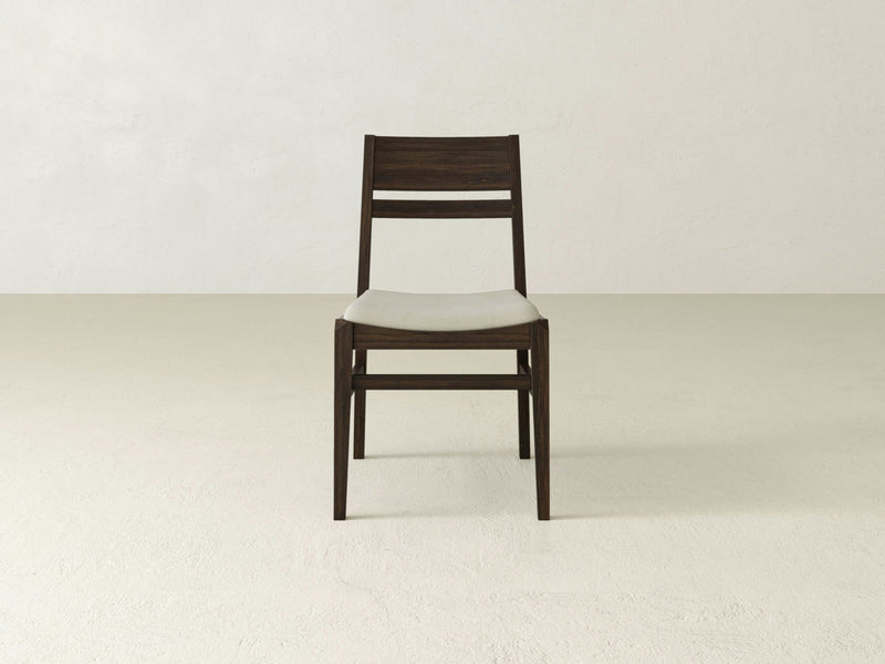 A Delilah Dining Chair - Tobacco by James+James with a simple, modern design sits centered in a minimalistic, light-colored room. The chair has a dark wooden frame and a light-colored padded seat, with horizontal slats on the backrest.