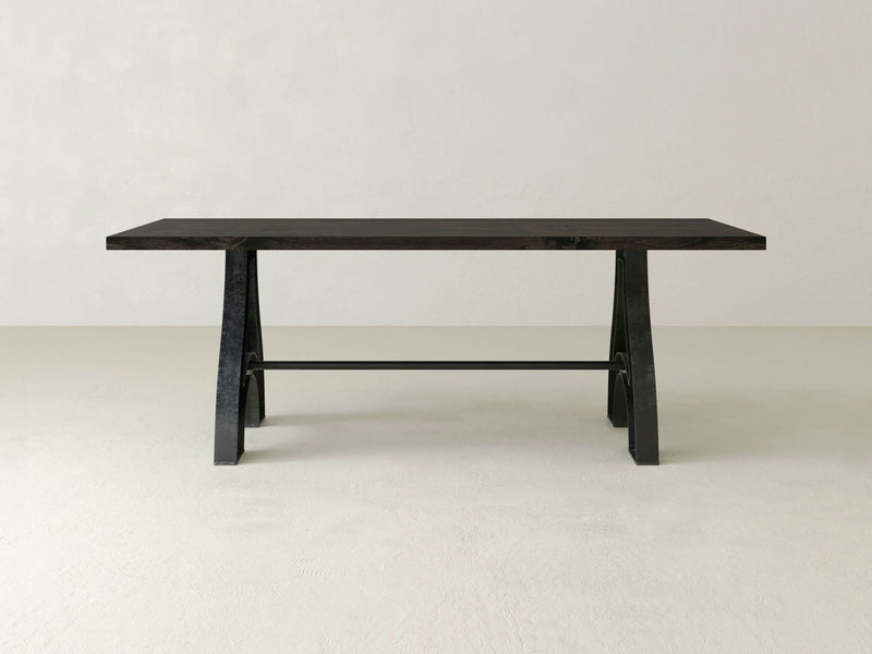 The Wishbone Dining Table - Charred Ember by James+James stands centered in an empty, white room. This minimalist dark wood table features a rectangular top with an A-frame base, showcasing a sleek and modern design. The clean lines and supportive crossbar between the legs enhance its contemporary aesthetic. Soft lighting creates a gentle shadow beneath the table.