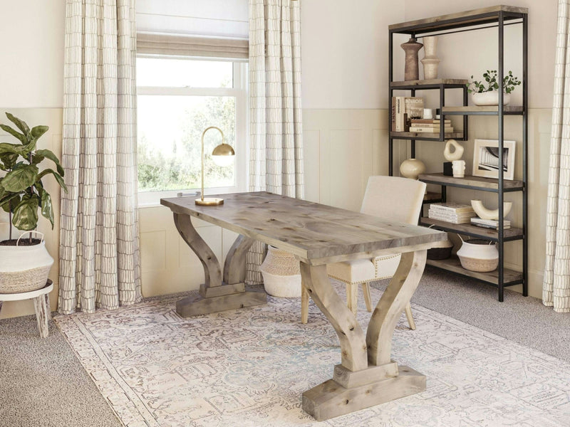 A home office featuring the Vera Desk - Barn Wood by James+James paired with an upholstered chair. The desk is adorned with a brass desk lamp. Against the wall stands a bookshelf decorated with various items. A window dressed in white curtains allows natural light to spill into the room, while a green potted plant occupies the corner.