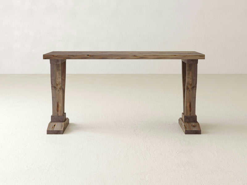 The Vera Sofa Table by James+James features a rustic design with a rectangular top and two sturdy, intricately crafted pedestal legs. Finished in barn wood, it beautifully showcases the natural grain and texture of the wood, set against a plain, light-colored background.