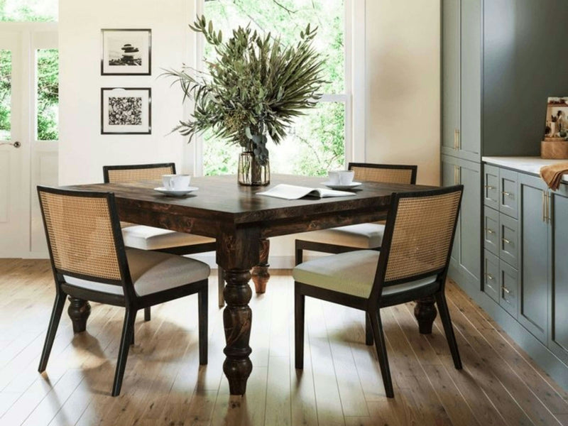 A cozy dining area features the Baluster Square Dining Table in Tobacco by James+James, surrounded by four cushioned chairs. A large glass vase filled with green branches serves as the table centerpiece. The light-filled room has wooden floors, framed wall art, and built-in shelving on one side.