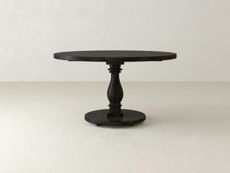 The Vivien Round Dining Table - Charred Ember by James+James stands on a light-colored floor against a plain, light wall. The table features a single turned column at the center, connecting the round tabletop to a circular base.