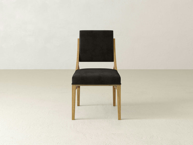 The Quinn Dining Chair - Harvest Wheat by James+James, featuring a minimalist wooden frame and dark upholstered seat and backrest, is placed in an empty room with light-colored walls and a light-colored floor. The chair boasts clean lines and a modern design.