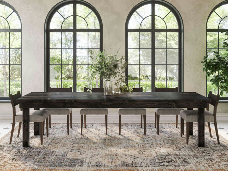 A modern dining room showcases the Farmhouse Dining Table - Charred Ember by James+James, paired with eight gray cushioned chairs. Large arched windows flood the room with natural light, while a lush green foliage centerpiece adorns the table, complemented by a patterned rug and a potted plant.