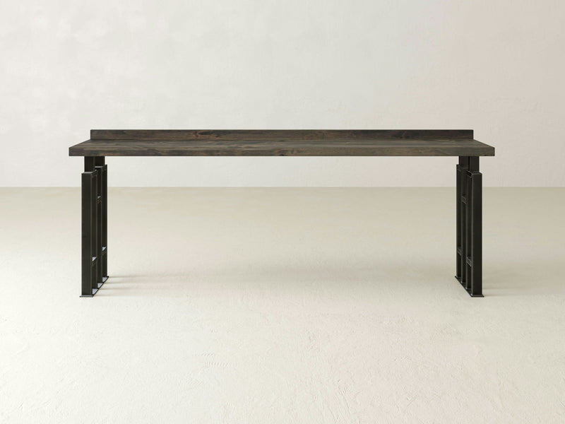 A Clark Sofa Table - Deep Grey by James+James stands on a light-colored floor against a plain, off-white background. The table features a dark wooden top with black metal legs, embodying a minimalist and modern design with clean lines and an industrial aesthetic.