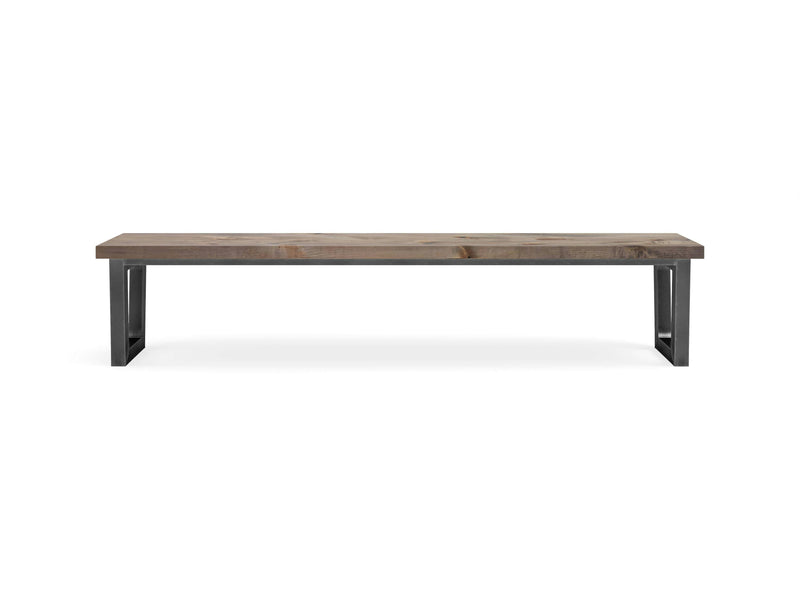 The Trapezoid Bench - Barn Wood by James+James features a natural finish and sleek, black metal legs forming its base. The design is minimalistic and modern, showcasing a long and sturdy wooden seat that beautifully highlights the wood grain. The bench is set against a plain white background.