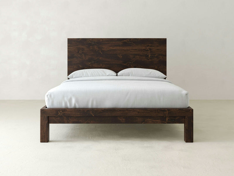 A minimalist bedroom scene featuring the Sawyer Bed in Tobacco by James+James, showcasing a dark wooden bed frame with a matching headboard. The bed is neatly made with light-colored linens and two pillows, set against a plain, light-colored wall. The overall aesthetic is clean and simple.