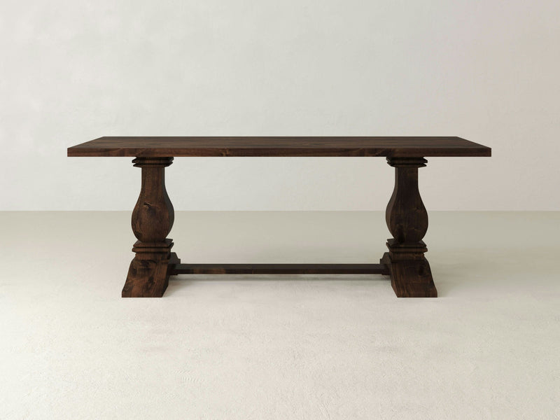 The James+James Heirloom Dining Table, a large handcrafted piece with a dark finish, is displayed against a plain white background. This exquisite dining table features a rectangular top and two ornate, baluster-style legs in a double pedestal design connected by a horizontal stretcher.