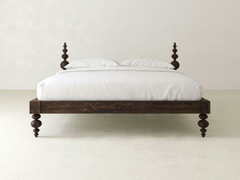 The Ophelia Bed - Tobacco by James+James, featuring intricately carved posts at each corner, stands on a white floor against a plain light-colored wall. The bed boasts a dark, rich finish and is neatly made with white linens, including two pillows.