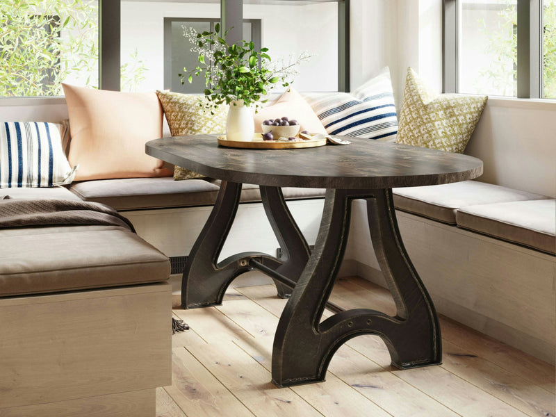 A cozy nook features the Wishbone Oval Dining Table - Tobacco by James+James, showcasing a unique pedestal base. It is surrounded by a built-in bench with cushions in various colors and patterns. A decorative tray with grapes and a green plant adorns the table, while large windows provide natural light.