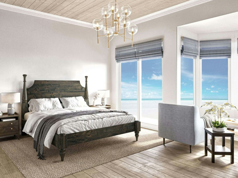 A serene bedroom featuring the Stella Bed in deep grey from James+James, complemented by light grey bedding and a matching grey armchair. Large windows offer a view of a bright blue sky and ocean. A beige rug, light wooden floor, and a modern chandelier complete the calming ambiance.