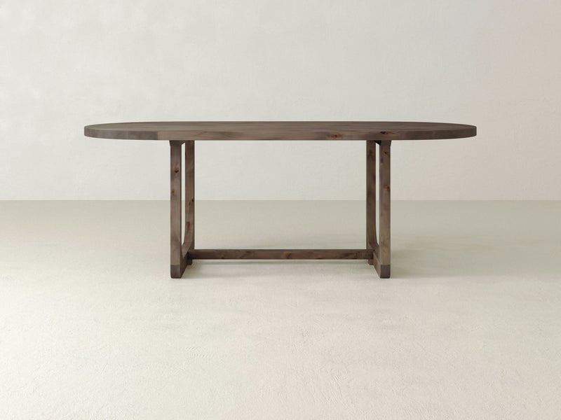 The Emersen Oval Dining Table - Barn Wood by James+James is featured. This table boasts a sleek, minimalist design with a smooth, dark brown surface. It stands on two sturdy, rectangular legs connected by a horizontal support. The backdrop is a plain, light-colored wall and floor.