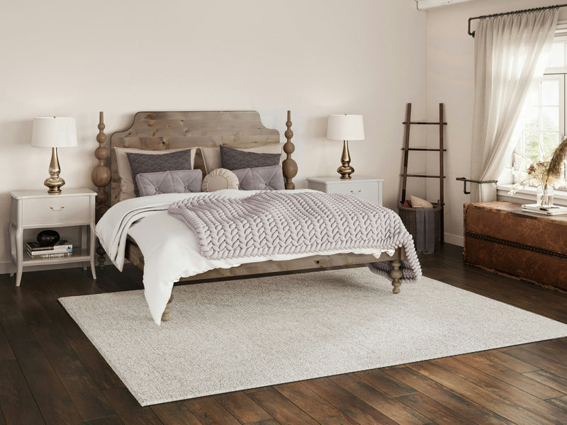 A cozy bedroom features the Opal Bed - Barn Wood by James+James, complemented by neutral bedding and matching nightstands with lamps. A ladder-style shelf leans against the wall, while a vintage trunk sits by the window. Large windows adorned with white curtains allow natural light to flood the room, highlighting the hardwood floor.