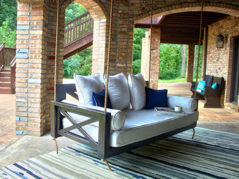 A cozy outdoor patio features a Finley Porch Swing - Charred Ember from James+James, adorned with white cushions and navy blue accent pillows. The swing is suspended under a brick archway, with a striped rug underneath. In the background, there's lush greenery and a wooden staircase leading up.