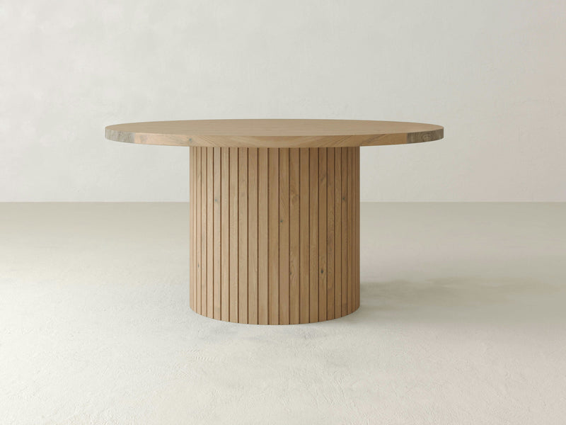 A round wooden Arthur Dining Table by James+James features a smooth, flat top and a cylindrical base adorned with vertical slats. Showcasing traditional craftsmanship, the table is set in a minimalist environment with a light, neutral-colored background.