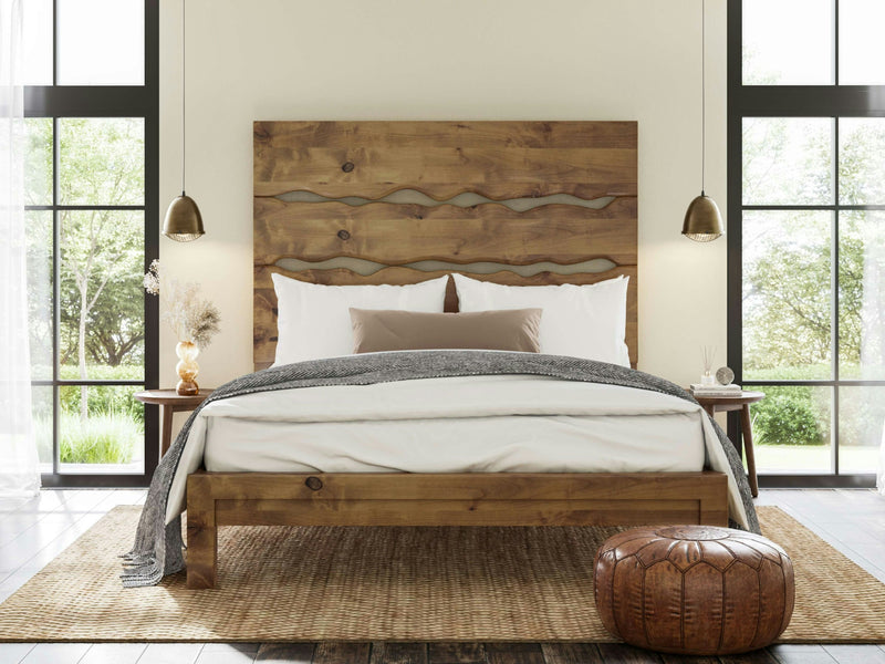 A cozy bedroom features the James+James Live Edge Bed in Harvest Wheat, distinguished by its unique wavy headboard. The bed is adorned with white and beige pillows and a gray throw blanket. Two pendant lights hang above wooden nightstands on either side. A round, brown ottoman and a jute rug enhance the rustic decor.