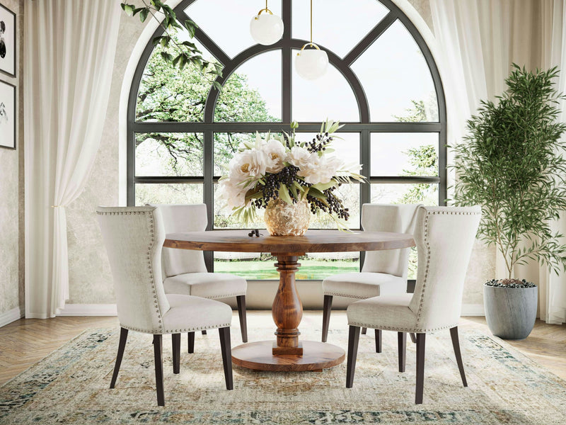 A dining room featuring the Vivien Round Dining Table - Tuscany by James+James, encircled by four beige upholstered dining chairs. A vase filled with white and dark purple flowers elegantly rests on the table. The background showcases large arched windows adorned with white curtains, offering a scenic view of lush trees. An indoor plant is placed on the right side of the room.