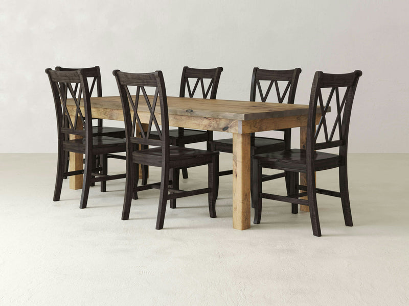 A rectangular wooden dining table with a rustic finish from the Farmhouse & Double X-Back Dining Set by James & James is surrounded by six Charred Ember Double X-Back Dining Chairs. Each chair features dark, crisscross backrests, and the set is arranged in a minimalistic room with white walls and a light-colored floor.