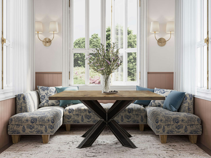 A well-lit dining area features the James+James Shiloh Square Dining Table in Harvest Wheat with an X-shaped base, surrounded by cushioned seating with floral-patterned upholstery. Two wall sconces with cream-colored lampshades are mounted on either side of the large windows, which offer a scenic outdoor view.