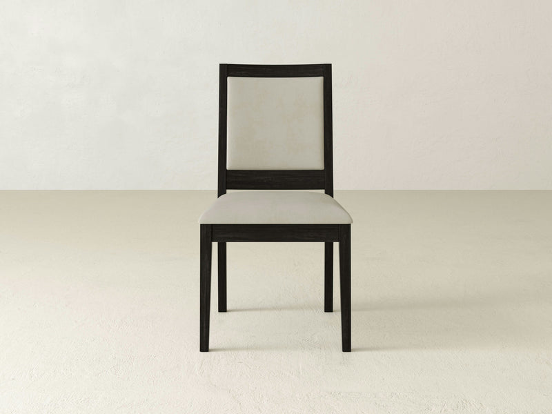 The Gina Side Dining Chair - Charred Ember by James+James, featuring a dark wooden frame and light-colored upholstered seat and backrest, is positioned centrally in a simple, neutral-toned room with no other furniture or decorations.