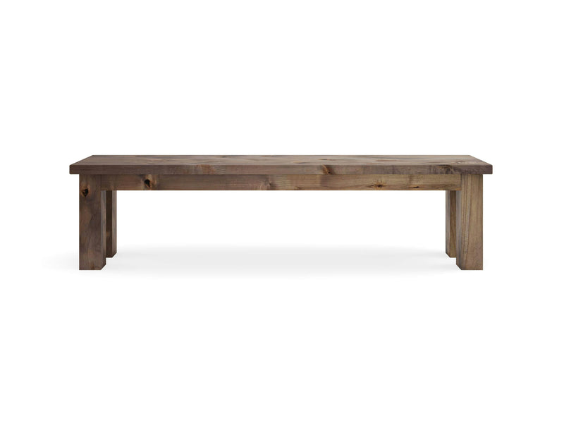 The Farmhouse Bench - Barn Wood by James+James features a simple, rustic design. This wooden bench boasts a rectangular seat supported by four sturdy legs. Its natural finish highlights the beautiful grain patterns and knots in the wood, offering a minimalistic and functional appearance.