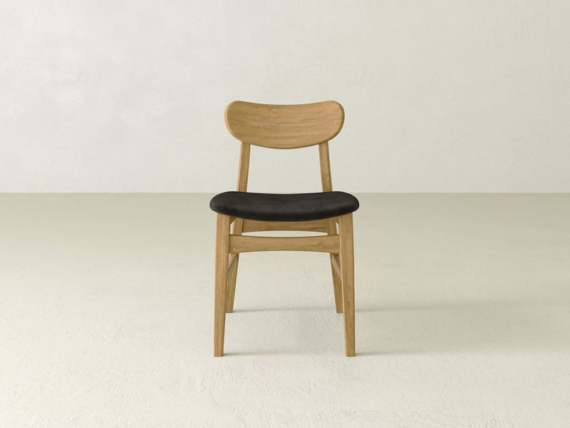 The Lynn Upholstered Dining Chair - Harvest Wheat by James+James is centered against a plain, light-colored background. This wooden chair features a curved backrest, black cushioned seat, minimalist design with slender legs, and a natural wood finish.