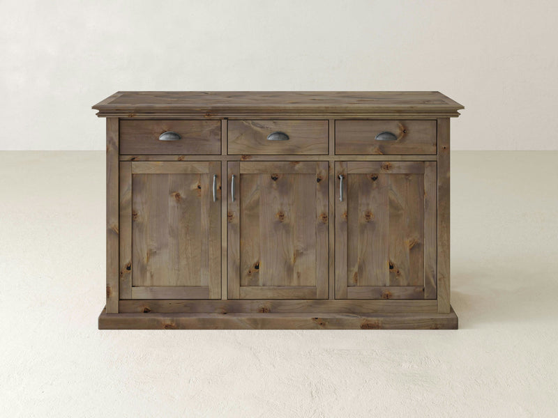 The Nora Buffet - Barn Wood by James+James is a rustic wooden sideboard with a light brown finish. It features three drawers with dark metal cup pulls and three cabinet doors with vertical panels and matching metal handles. The piece has a simple, sturdy design and sits on a flat base.