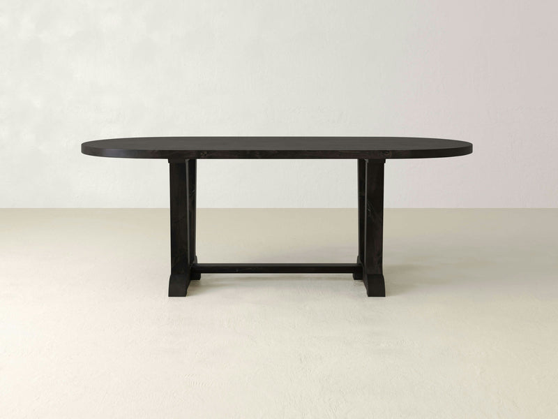 A simple, modern Thaden Oval Dining Table - Charred Ember from James+James, featuring a dark-colored, flat surface and sturdy legs. The table is centrally placed against a plain, light-colored background.