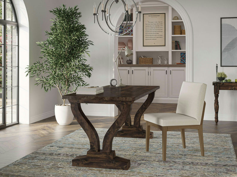 A bright, airy room with wooden flooring features the Vera Desk in Tobacco by James+James, showcasing its elegant curved legs. The space is adorned with a beige cushioned chair, a large potted plant, and a cabinet with shelves holding books and decor. A modern chandelier hangs from the ceiling, while an arched window allows natural light to fill the room.