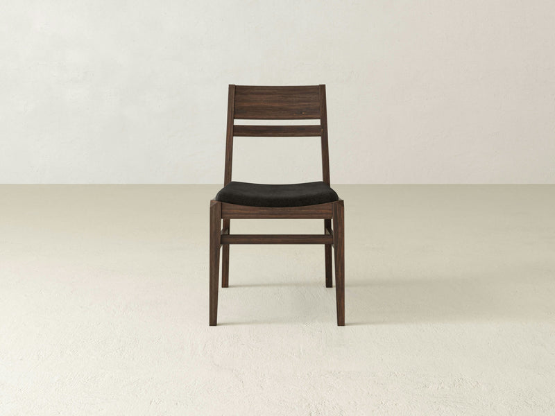 The Delilah Dining Chair - Tobacco by James+James, featuring a wooden frame with a dark finish and a black cushioned seat, is positioned against a plain light-colored background. The chair showcases a slatted backrest and simple, clean lines.