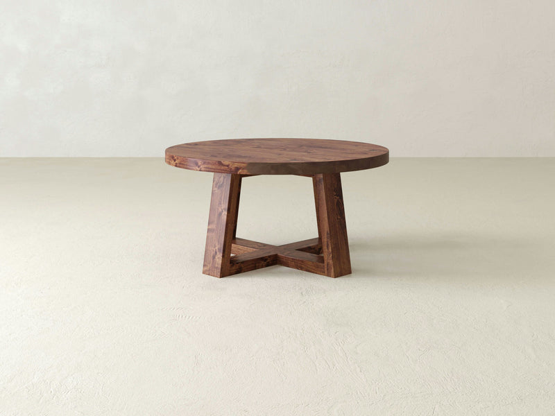 The Rae Coffee Table - Tuscany by James+James, a round wooden table with a rustic finish, stands on a light-colored floor. The table features a thick top and a sturdy, angular base with a unique cross-brace design. The background is a plain, muted wall.