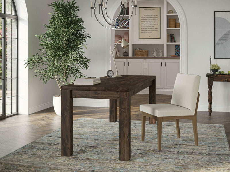 A modern home office featuring the Jonathan Desk - Tobacco by James+James and a beige upholstered chair. The room includes a lush potted plant, a large window, built-in white shelving with books and décor, a chandelier, and a patterned rug under the desk and chair.