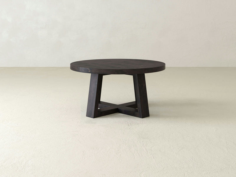 A Rae Coffee Table - Charred Ember from James+James is placed in a minimalist room with light-colored walls and floor. This round wooden table features a sturdy, geometrically designed base with intersecting legs and has a dark finish.