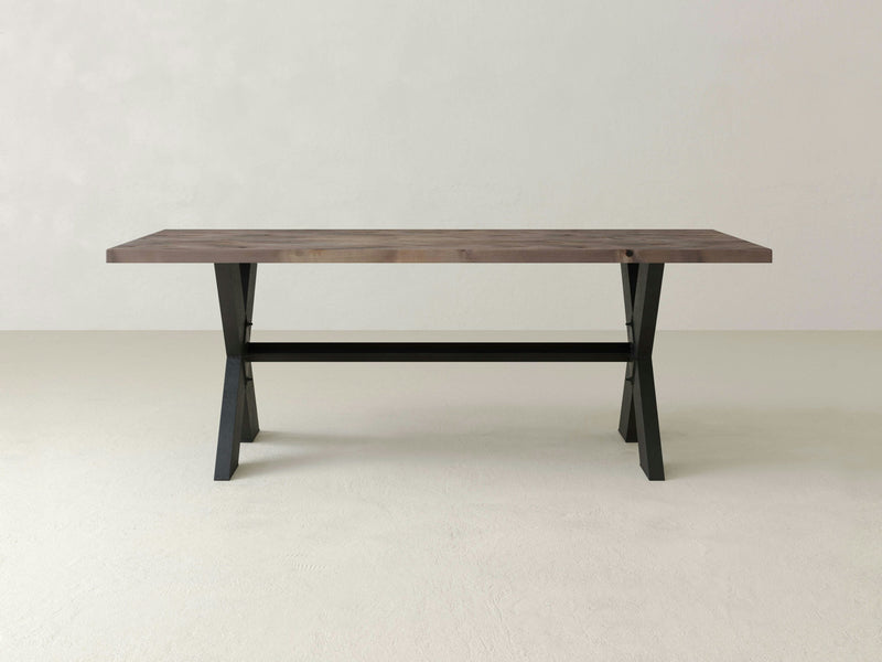 The X-Base Dining Table - Barn Wood by James+James features a minimalist wooden top with a rectangular shape and black metal legs. The design is simple and modern, with an open space beneath the table and a horizontal support bar connecting the legs. The background is a plain, off-white wall and floor.