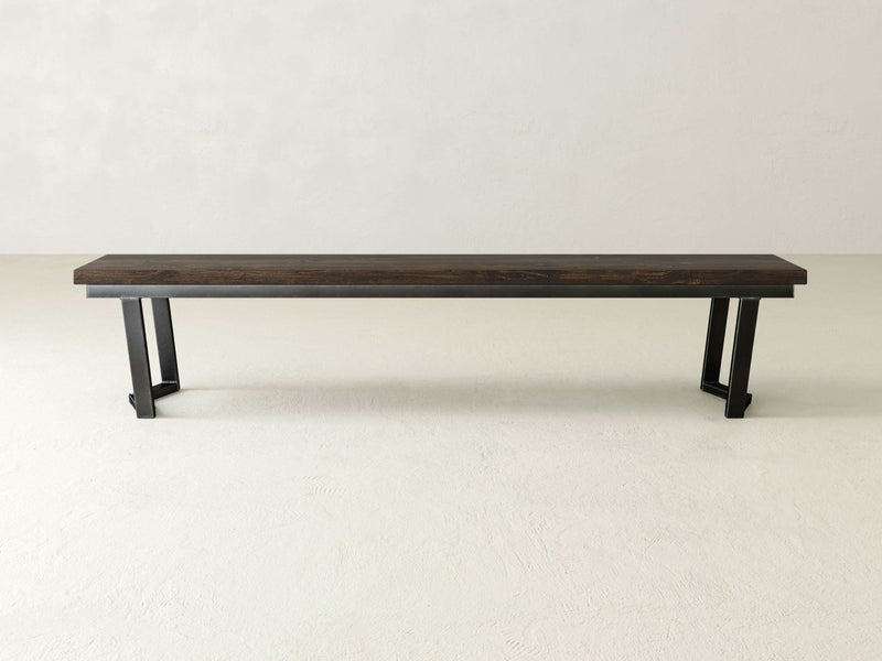 The Arkwright Bench - Tobacco by James+James features a simple design with a dark brown seat and black metal legs. It is positioned on a light-colored floor against a light wall, embodying a minimalist and contemporary aesthetic with clean lines and a sturdy appearance.