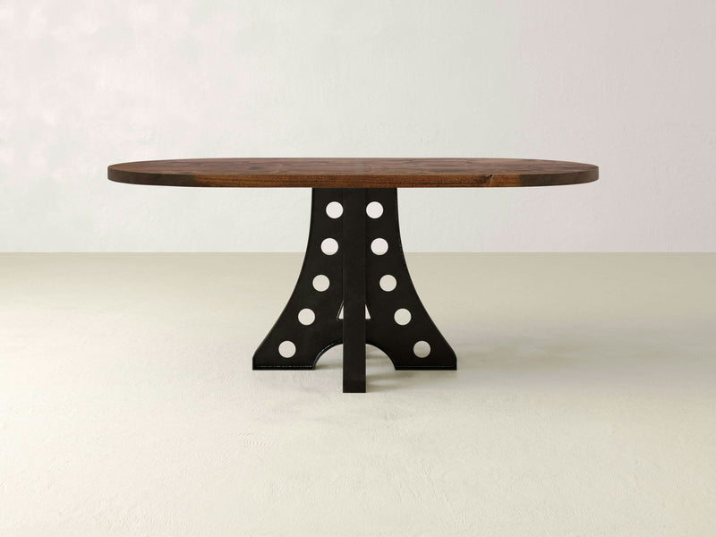 The Amelia Oval Dining Table - Tuscany by James+James showcases a sophisticated dark-finished wooden top and a uniquely designed black metal base with circular cutout patterns. The table is elegantly displayed against a plain, light-colored background.