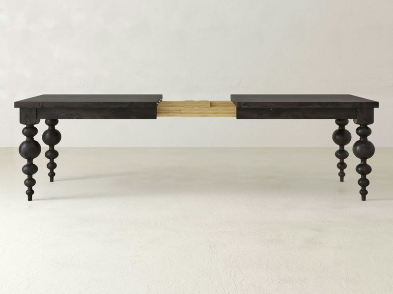 The Olivia Expandable Dining Table - Charred Ember by James+James features a dark brown wooden finish with intricately carved, turned legs. The table is extended in the center, highlighting its extension mechanism, which allows it to be lengthened. The background showcases a plain, light-colored wall and floor.