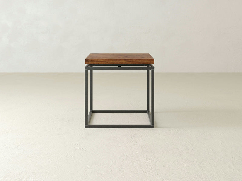 The Floating Top End Table - Tuscany by James+James, featuring a minimalist wooden tabletop and a sleek black metal frame, sits on a plain, light-colored floor against a light gray wall. This end table showcases a simple, modern design.