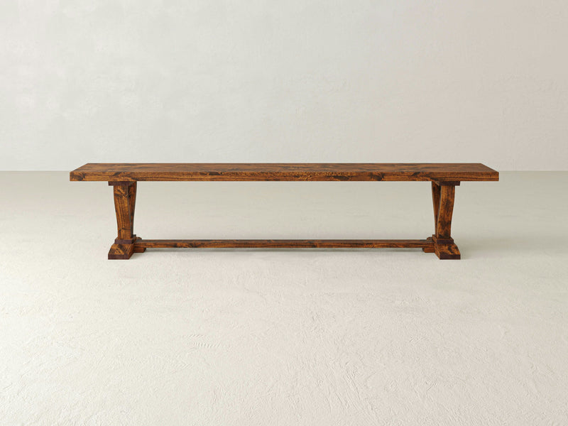 The Vera Bench - Tuscany from James+James is a rustic wooden bench with a simple yet sturdy design, featuring a long flat seat and two thick support legs connected by a horizontal brace. The bench is set against a plain white background.