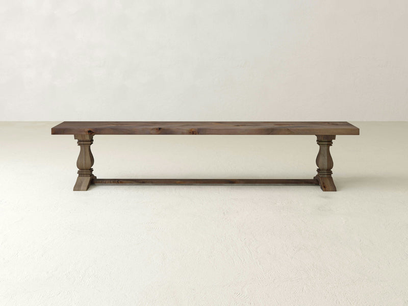 The Heirloom Bench - Barn Wood by James+James features a flat, rectangular seat and two sturdy, turned legs connected by a thick horizontal support beam. It is positioned on a light-colored floor against a plain, light gray background.