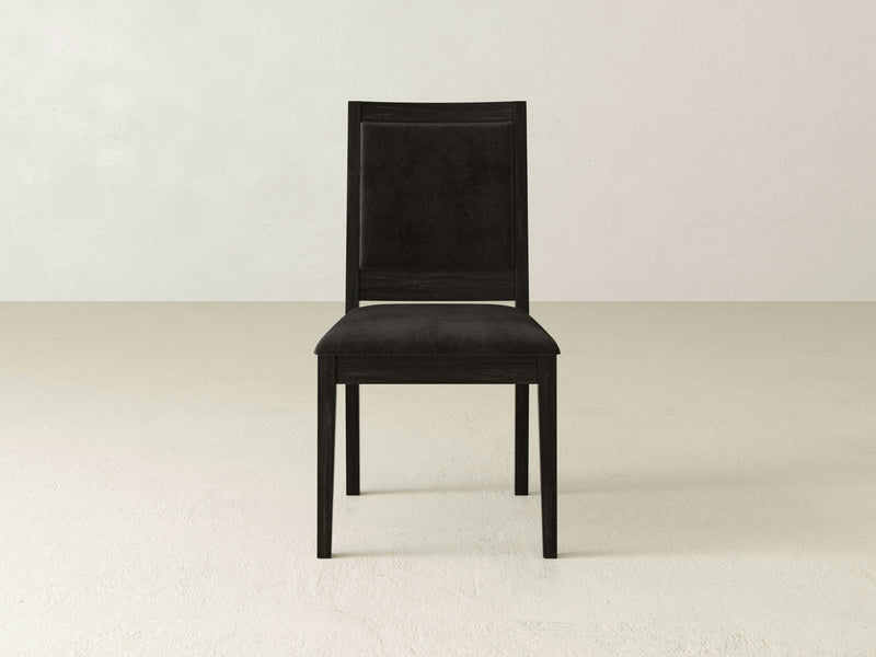 A single Gina Side Dining Chair - Charred Ember from James+James, featuring a black wooden frame with a cushioned seat and backrest, stands on a light-colored, textured floor. The minimalist design boasts simple, straight lines and a smooth finish. The background is a plain, off-white wall.