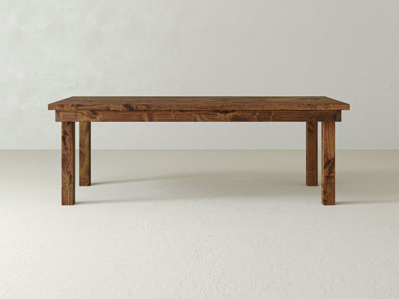 The James+James Farmhouse Dining Table - Tuscany, designed with a rectangular shape and four sturdy legs, stands against a plain, light-colored background. The table boasts a smooth finish and natural wood grain, showcasing a simple yet rustic design.