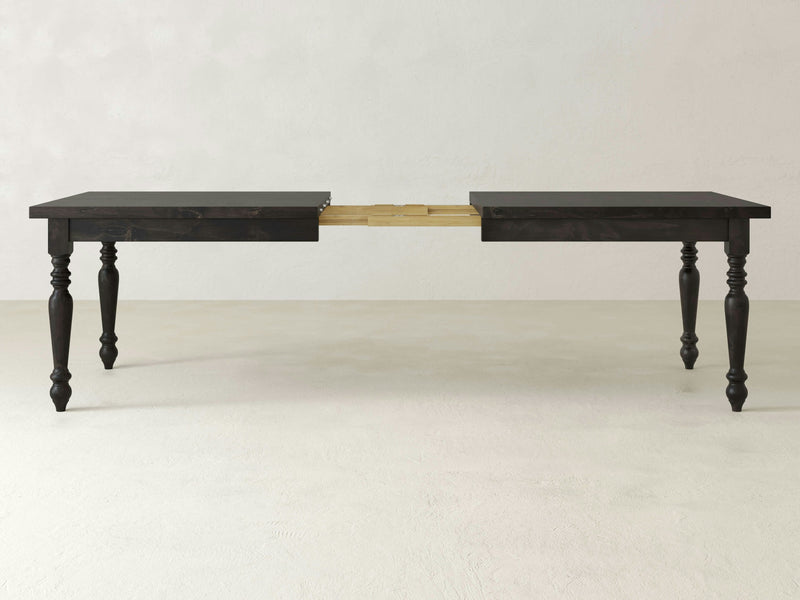 The French Country Expandable Dining Table - Charred Ember by James+James features a dark wooden design with ornate, carved legs. It includes an extendable central section for length adjustment, and its rectangular tabletop is highlighted against a plain, light background.