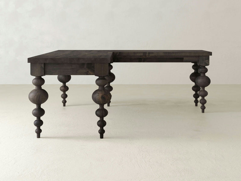 The Olivia Corner Desk by James+James is a deep grey L-shaped table featuring distinctively carved, spherical and bulbous legs. Its symmetrical design exudes a rustic charm, showcasing intricate craftsmanship with a bohemian flair. Photographed against a plain white background, this piece stands out as an elegant addition to any space.