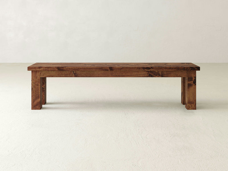 A close-up of the Farmhouse Bench - Tuscany by James+James reveals a wooden surface with a prominent knot in the center. The wood has a rich, warm brown color with visible grain patterns running horizontally. The knot, featuring a dark, irregular shape, adds texture and character to this beautifully crafted piece.