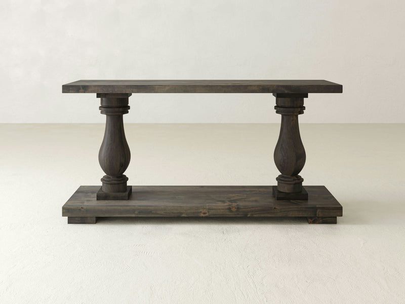 The Vivien Sofa Table by James+James is a rustic wooden console featuring a dark finish that seamlessly blends form and function. Its design includes a flat top supported by two ornately carved, baluster-style legs connected to a thick rectangular base, with its robust construction standing out against the simple, neutral background.