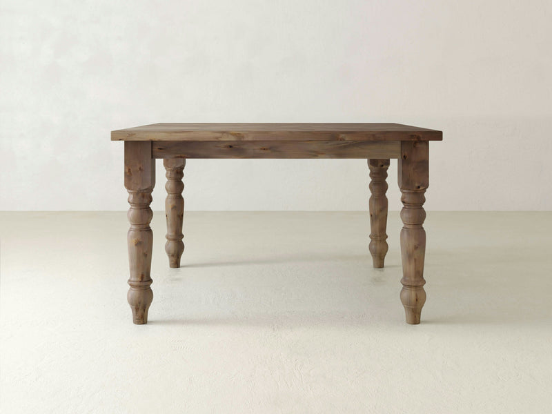 A rustic Baluster Square Dining Table - Barn Wood by James+James, featuring a plain rectangular top and ornate, carved legs, stands on a light-colored floor against a white wall. The table has a natural wood finish, showcasing its grain and texture.